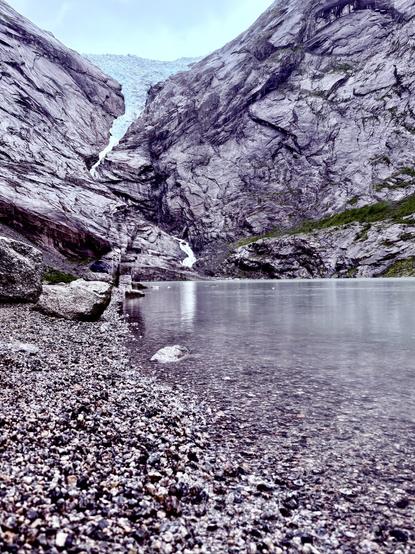 Two mountains with a glacier between them. In the foreground is a lake and a pebble beach.