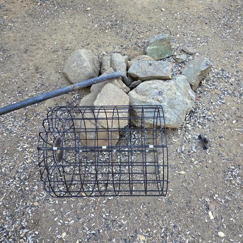 A black metal pole is bent at a ninety-degree angle over large stones that were anchoring it. The black wire basket of the birdfeeder lies on barren ground covered with thousands of sunflower seed shells.