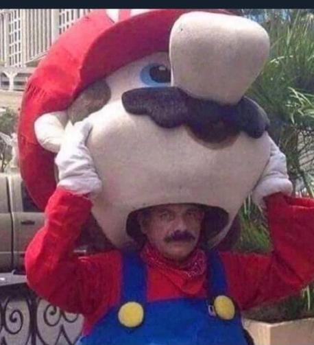Une personne en costume de Super Mario (homme portant une salopette bleue sur un maillot à manches longues rouge, une casquette rouge ornée d'un M et une volumineuse moustache), en train d'enlever la tête du costume, révélant un homme portant une volumineuse moustache similaire à celle du costume