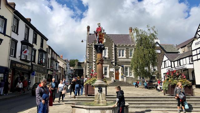The photo depicts a lively street scene in a charming town square, likely in Wales, given the flags and architectural style. At the center is a tall statue of a crowned figure holding a shield with a red lion emblem, standing atop a decorative column. At the base of the column is a fountain with water spouting from a sculpted lion's head. Surrounding the square are traditional buildings with white facades, black timber framing, and stone structures. Shops like "NEW CHOICE" and "T E R A J" are visible on the left, alongside a restaurant named "Alfredo Restaurant" on the right. The area is bustling with people walking, shopping, and relaxing, with flower arrangements adding vibrant color. The sky is partly cloudy, casting a bright, cheerful light over the scene.