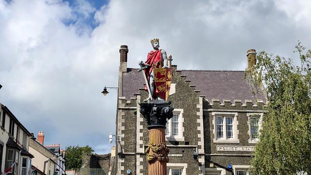 This picture shows a statue of a crowned figure, likely a king, standing atop a tall ornate column. The figure is dressed in a red cloak, holding a sword in one hand and a shield featuring a lion emblem in the other. The column is detailed with decorative carvings. In the background, there is an old stone building with distinct Gothic-style architecture, labeled "POLICE STATION." The structure has a dark, textured façade with white stone trim around the windows and along the edges. There are street lamps, a sign reading "TUDOR SQUARE," and some surrounding buildings, including visible rooftops and chimneys. A tree partially frames the right side of the image under a sky filled with clouds and patches of blue.