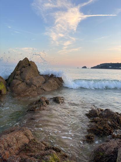 Waves crash against rugged rocks on a peaceful shoreline under a sky with wispy clouds at sunset.