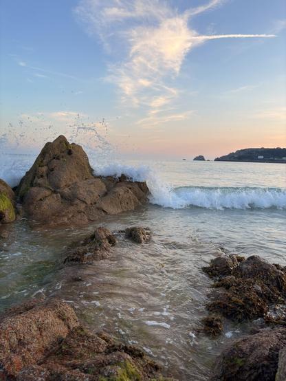 Waves crash against rugged rocks on a peaceful shoreline under a sky with wispy clouds at sunset.