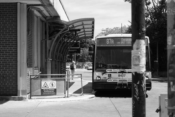 Looking from Broadview Avenue into the south bus platform. At right, the 87A Cosburn bus sits face on to the camera. Between the camera and the bus, a utility pole with a tattered poster runs from top to bottom of the frame. At left is the brick facade of the station and the curved overhang of the roof over the platform. Framed by that curve, one lone passenger stands, looking away from the camera.