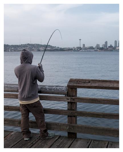Person fishing on a pier, with the Seattle skyline in the background.