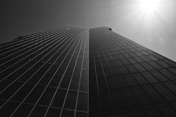 Dramatic upward view of a modern glass skyscraper under a bright sunburst in the top right corner. The building’s reflective facade and sharp vertical lines create a sense of height and architectural symmetry.