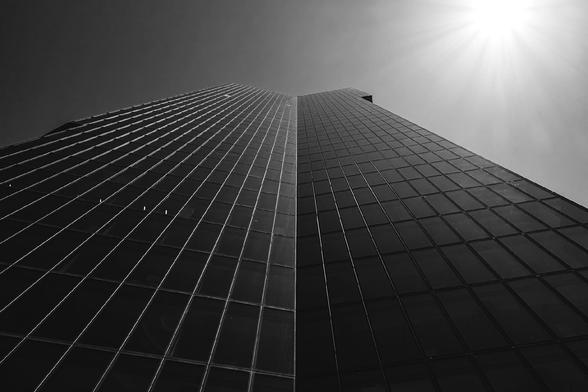 Dramatic upward view of a modern glass skyscraper under a bright sunburst in the top right corner. The building’s reflective facade and sharp vertical lines create a sense of height and architectural symmetry.