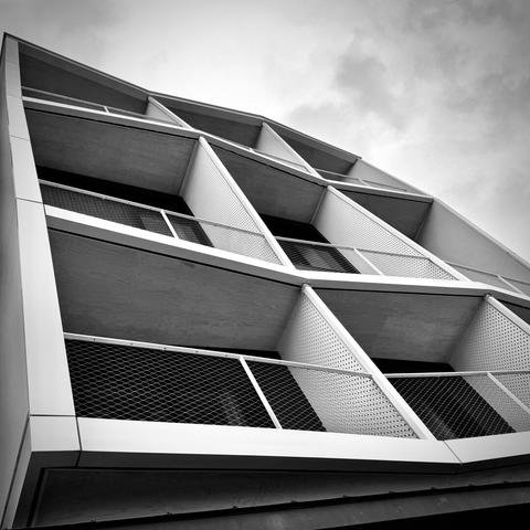 A dutch angle views a nine-by-nine-block neo-expressionist apartment building with balconies of numerous different dimensions under a cloudy sky above.