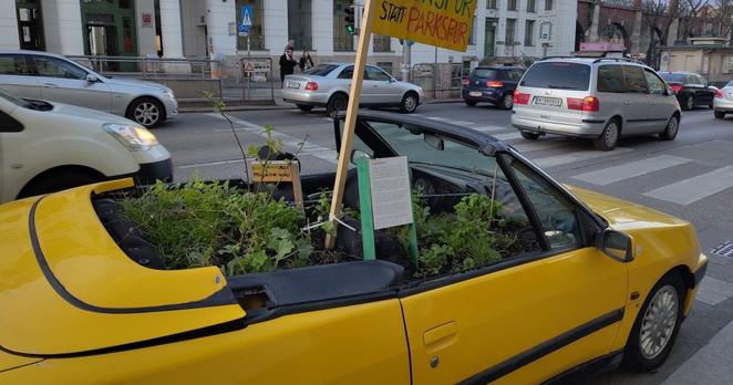 Der Vordergrund des Fotos zeigt ein gelbes Cabrio mit offenem Verdeck von der Seite. Es ist mit Erde und Pflanzen gefüllt, auf einem darin befestigten Schild steht handgeschrieben der Slogan „Grünspur statt Parkspur“. Das Cabrio steht am rechten Rand einer mehrspurigen Straße. Daneben und davor sind fahrende Autos zu sehen, im Hintergrund eine U-Bahn-Station und zwei Erwachsene mit einem kleinen Kind, die darauf warten, die Straße überqueren zu können.