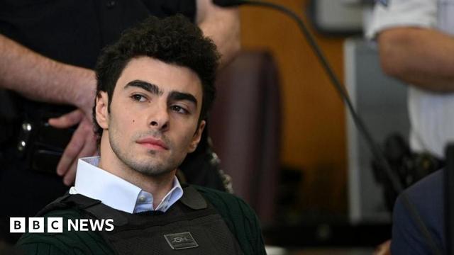 Luigi Mangione, wearing a collared shirt, a green sweater and a bullet-proof vest, sits in court. 