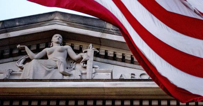 An American flag waves over the roof of a building with a woman representing justice, a sword in one hand and another weighing a feather.