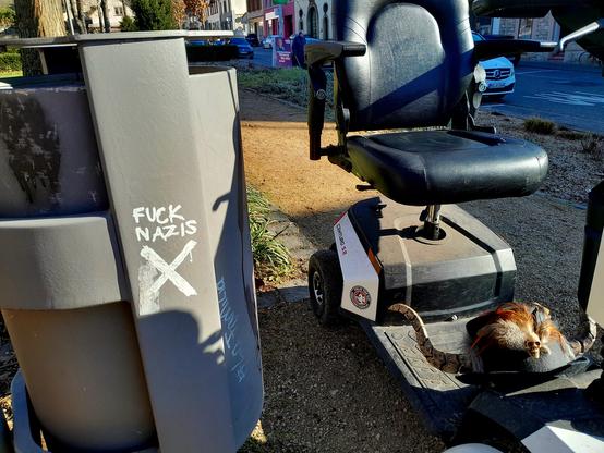The scoot-chair is parked next to a trash can emblazoned with FUCK NAZIS, and the Shitter logo, which is a sans-serif swastika. 
Notice the hat on the floorboard.