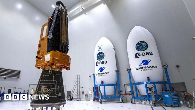 The biomass satellite sits inside an all white warehouse. It has a black cone-like base, an orange l-shaped top that has the look of foil, with the antenna attached and furled. To the left of the satellite sits two shells which will hold the satellite, they are white elongated cones with "ESA" written on them, and held in blue scaffolding trolleys. 