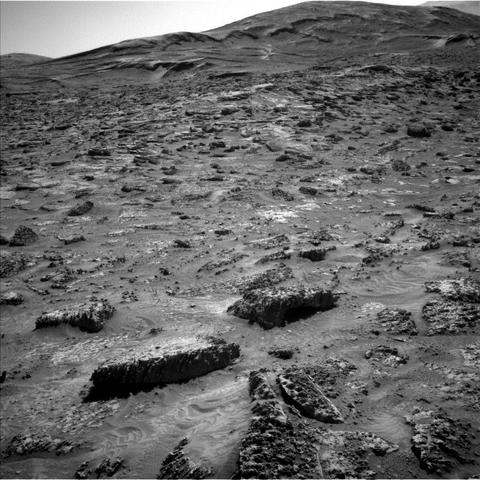 A grayscale photograph of the Martian surface from the Curiosity rover shows a field of rough and jagged rocks covering the surface from the foreground extending to hills in the background, all medium to dark gray. The hills appear smoother overall, but have horizontal folds on their sides leading up to their peaks.