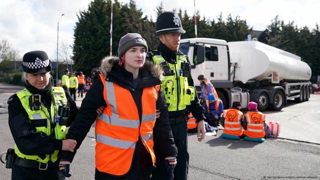 A young women in an orange hi-vis jacket is held by two police officiers, an oil tanker in teh background