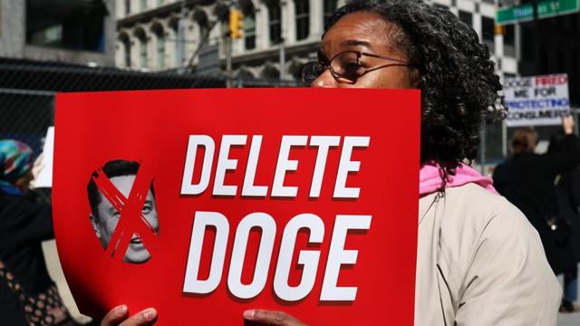 A person holds a sign that says "Delete DOGE" and has a picture of Elon Musk's face with an X over it.