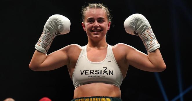 Georgia O'Connor smiles and holds both of her hands, in boxing gloves, up near her ears as she celebrates victory in the ring