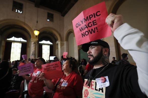Los Angeles residents hold signs reading "OLYMPIC WAGE NOW" during an indoor rally