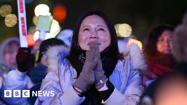 A woman wearing grey gloves stands with her hands clasped together against a night time backdrop