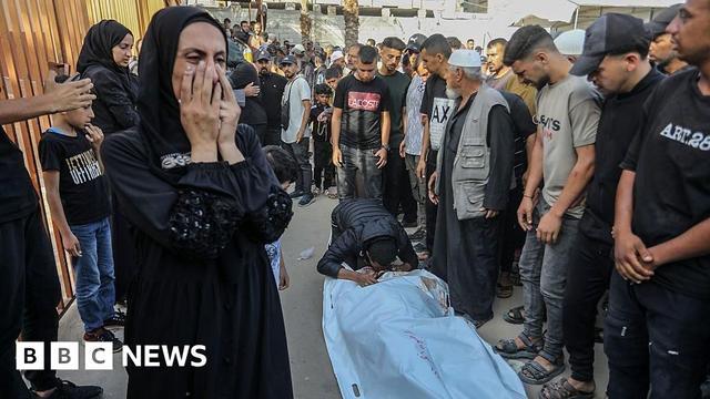 A body bag lies on a ground while a man kneels to kiss it. Others gather round while a woman covers her face and cries.