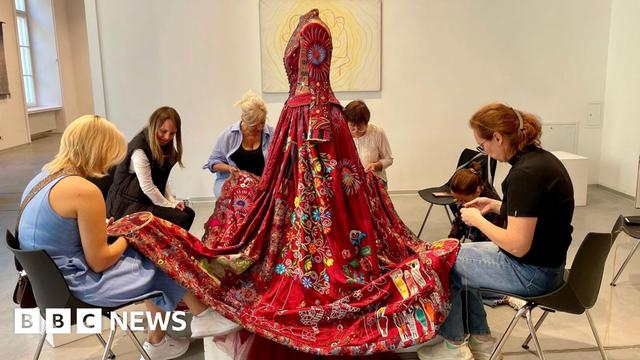 A red dress on a mannequin with women sat on chairs around the dress and embroidering it.