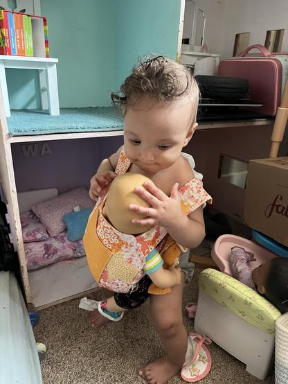 A 9-month-old with curly hair is playing with a baby doll, which is secured in a colorful carrier. The background includes a dollhouse with books, a stack of items, and another doll in a mini chair.