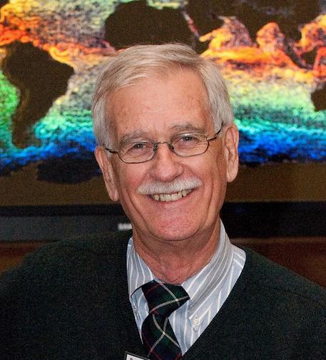 A headshot of a man smiling in front of a screen showing colorful data about Earth's oceans. he has grey hair, glasses and a moustache.