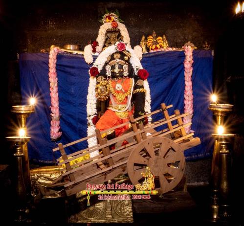 Idol of Hindu deity Krishna in red and many gold ornaments. A small old-timey wooden cart placed sideways in front of Krishna. Krishna's right leg bent slightly to as if kicking the cart. Thick white flower garland over Krishna and then flowing down on either side of Krishna. Blue cloth background. A garland of pink Lotus flowing down on either end of the background. Four decorative brass pedestal lamps surround Krishna and the cart. The lamps are lit.