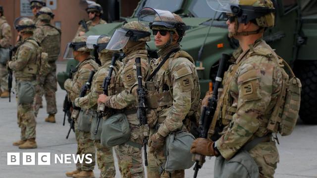 National Guard soldiers, wearing camouflage uniforms and helmets with visors lifted up stationed at the Edward R Roybal federal building in Los Angeles, with more soldiers and police in the background.