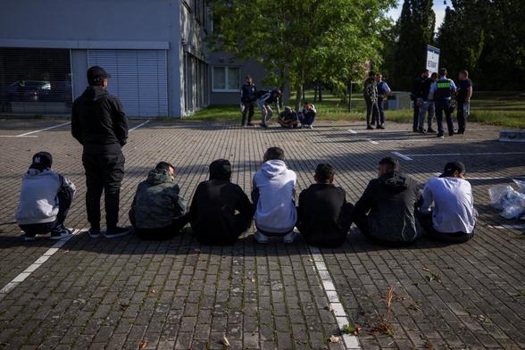 Rückansicht von Menschen, die auf einem Parkplatz in der Reihe teilweise hocken oder sitzen. Im Hintergrund ist ein Polizist erkennen, der sich mit anderen Menschen unterhält