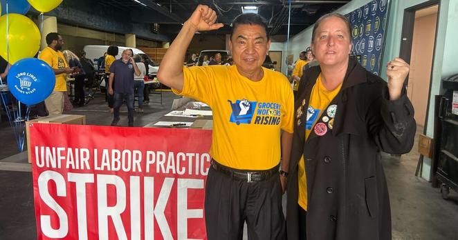 A medium-skin tone person wearing yellow t-shirt stands next two a light-skin tone person wearing a black coat adorned with pins, on top of a yellow t-shirt. The two people stand, with a fist in the air, in front of a bright red sign that says "Unfair Labor Practice Strike" in bold, white letters. 
