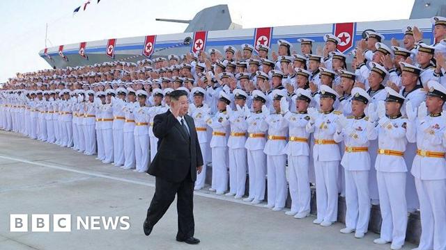 Rows of soldiers in white uniforms and white caps cheer as Kim Jong Un walk in front of them, wearing a black suit and raising his hand. Behind them is a massive grey ship.