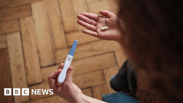 A woman holds a pregnancy test in one hand and a pill in the other