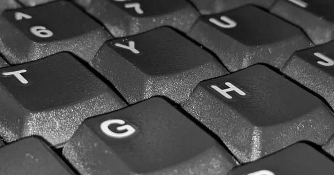 Close-up of a computer keyboard, seen diagonally from above. 6 keys are fully visible, with partially visible keys around the edge. They're dark gray with white markings, and close enough that you can see the texture on the sides of the keys and an outline around each letter suggesting that they were attached as stickers.