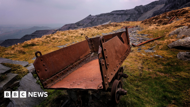 An old rusted quarry cart sits on a quarry hillside with a single track remaining. In the distance is grey slate quarrying terraces cut into the mountainside