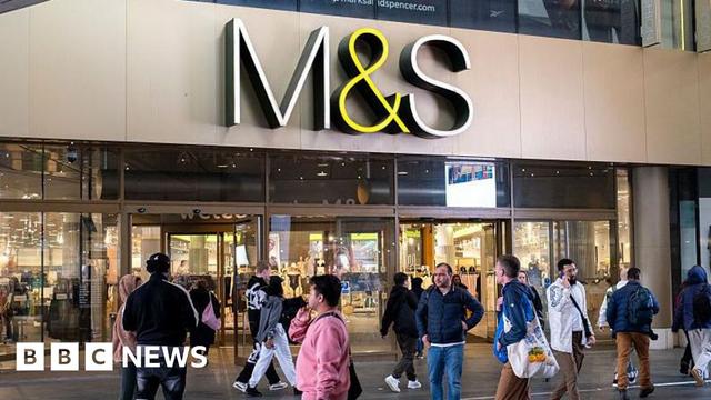 A crowd of people walk outside of a large M&S store with an M&S sign hanging overhead A crowd of people walk outside of a large M&S store with an M&S sign hanging overhead