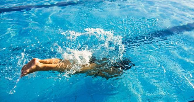 Ein Mensch taucht kopfüber in ein hellblaues Freibadbecken. Wasser spritzt auf, die Beine ragen noch aus dem Wasser. Die Sonne scheint auf der Wasseroberfläche.