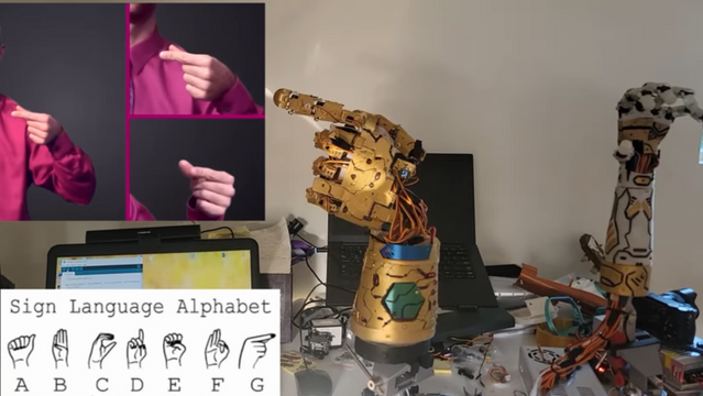A golden robotic hand is shown in the main picture performing the sign for the letter "g": pointing to the left, with all fingers except for the index finger curled. In the top left of the image, a human hand is shown imitating this position. A golden robotic hand is shown in the main picture performing the sign for the letter "g": pointing to the left, with all fingers except for the index finger curled. In the top left of the image, a human hand is shown imitating this position.