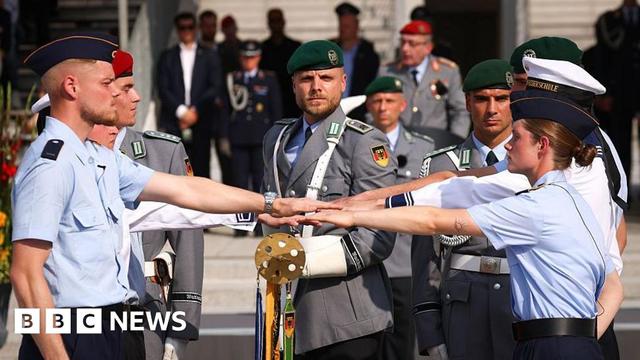 German army recruits take an oath, dressed in light blue shirts and dark blue berets as officers look on