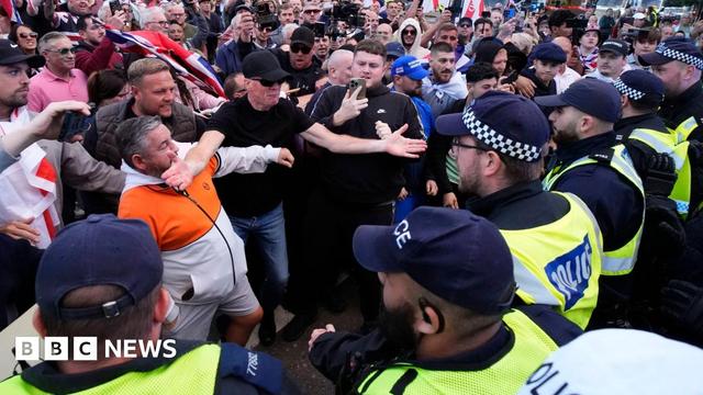 A line of police officers on the right of the picture are faced with a group of protesters, several of who are carrying flags and signs. One man stands holding his phone towards the confrontation 