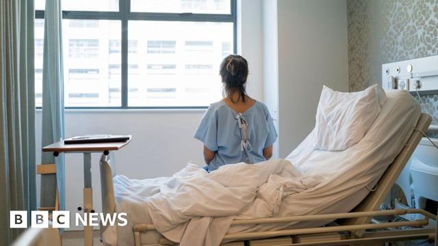 An anonymous woman sitting in an upright hospital bed. She is looking out of a window and her back is facing the camera. She is wearing a blue hospital gown.
