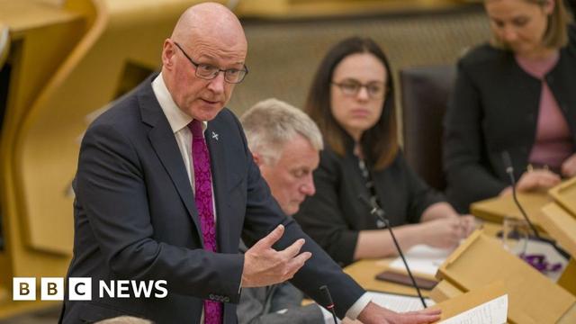 John Swinney, a bald man with glasses, speaks at a podium in the Scottish Parliament. He is wearing a dark suit, white shirt and purple tie, with his right hand gesticulating in front of his body. There are MSPs sat on benches behind him.