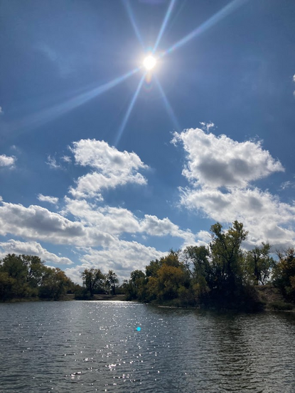 Bright sun with small reflective ripples on the surface of a lake surrounded by trees. A few clouds. 
