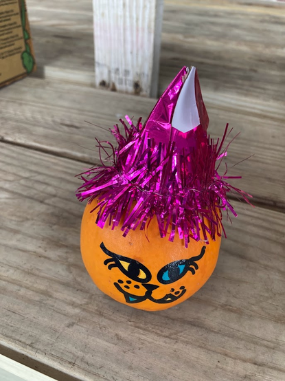 mini pumpkin with a cat face wearing a shiny pink tasseled hat. Background is wooden boards.