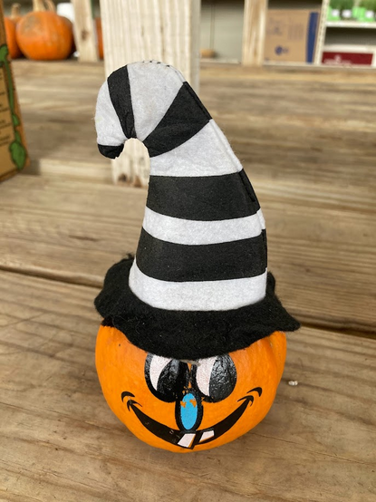painted face mini pumpkin wearing an oversized black and white felt hat. Background of wooden boards and other pumpkins. 