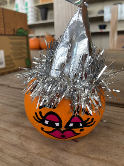 Painted pumpkin face with a shiny aluminum foil hat. A box, shelves and multiple other pumpkins are clustered together in the background. 