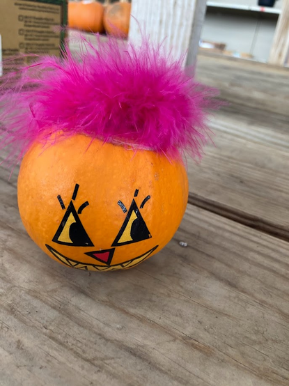 A mini pumpkin with a painted face wearing pink feathers as hair. 