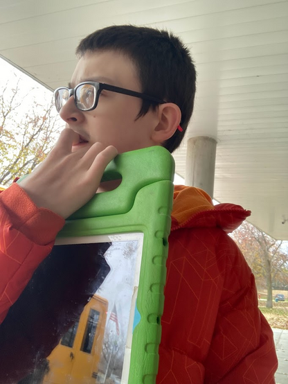 Jake a bit nervous about the field trip. We are standing outside the entrance, just about to go in. He does not realize yet that it's completely designed to optimize accessibility and comfort to him. He's chewing on one of his fingers while carrying his communication device as he cannot speak. He's looking off into the distance. He cannot see them but there were a large amount of geese in a small parkway but they were making a lot of noise. The communication device is an ipad with a green foam cover. There is a reflection of a school bus and flag visible. 