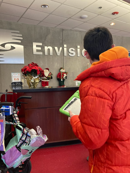 The front entryway, Jake is slowly checking out the Christmas decorations. One of his classmates wheelchair is also pictured. We were a group with four wheelchairs.

The wall is gray cement with bold letters along with a symbol art of two hands around a circle, Envision. There were poinsettias on the front desk. Although he's wearing his orange coat, it was at this point he decided he wanted to take it off and get comfortable. Bright red floor. 