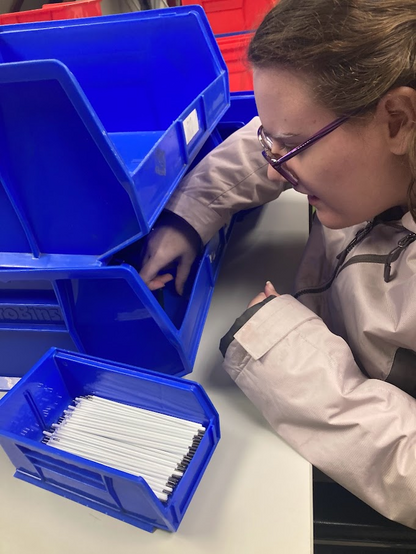 In addition to the factory there is an office setting for blind and IDD who may need a more sheltered environment. Here small tasks are performed with as much freedom as possible. Julia absolutely loved being able to sort and build things. Here she is pictured at a workstation. Two large plastic bins and a smaller one containing ink refills. They make pens as one of the products. Unlike Jake, she preferred to keep her coat on. She has one hand inside a plastic bin and is working on pulling out the pieces. 